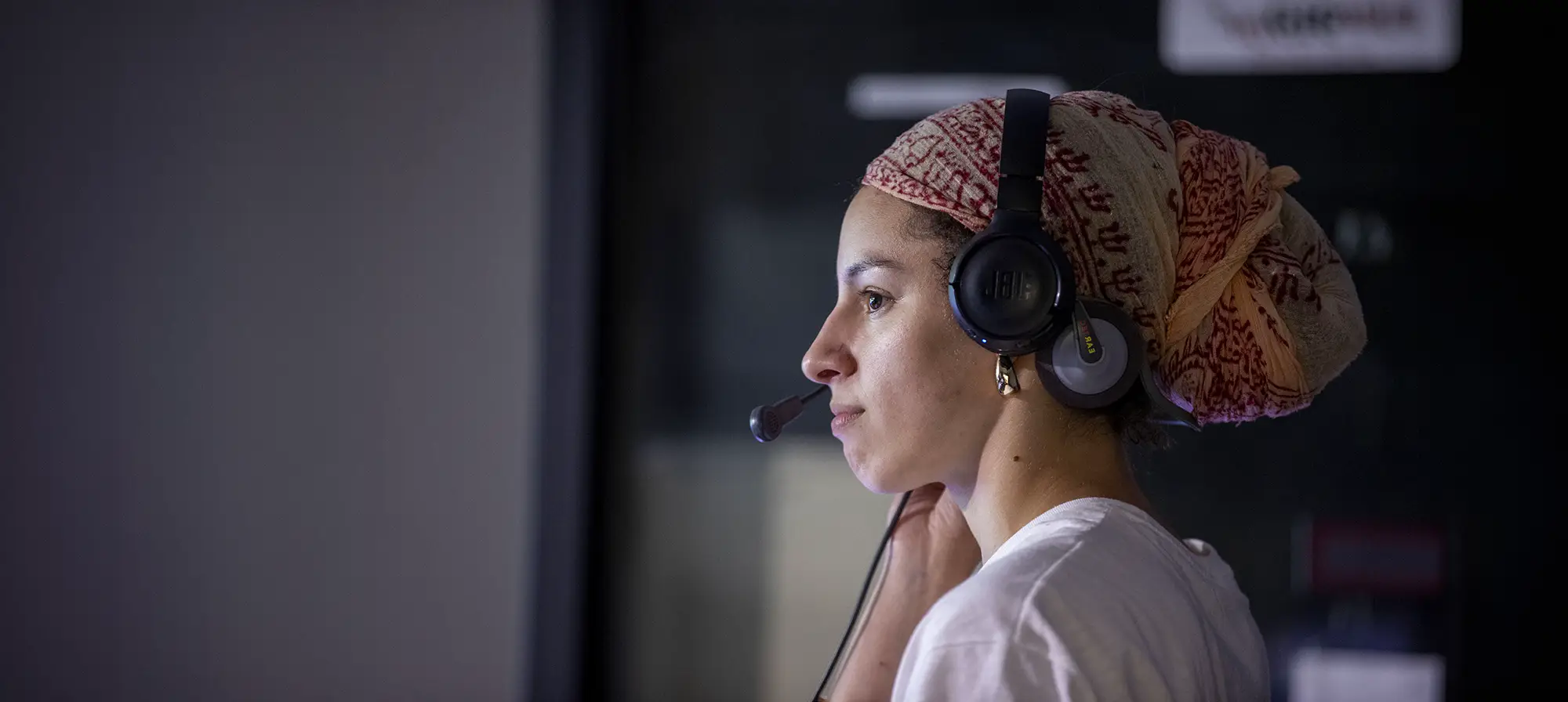 A student wearing a headset with a microphone looks at a monitor in a dimly lit broadcast or media production studio.
