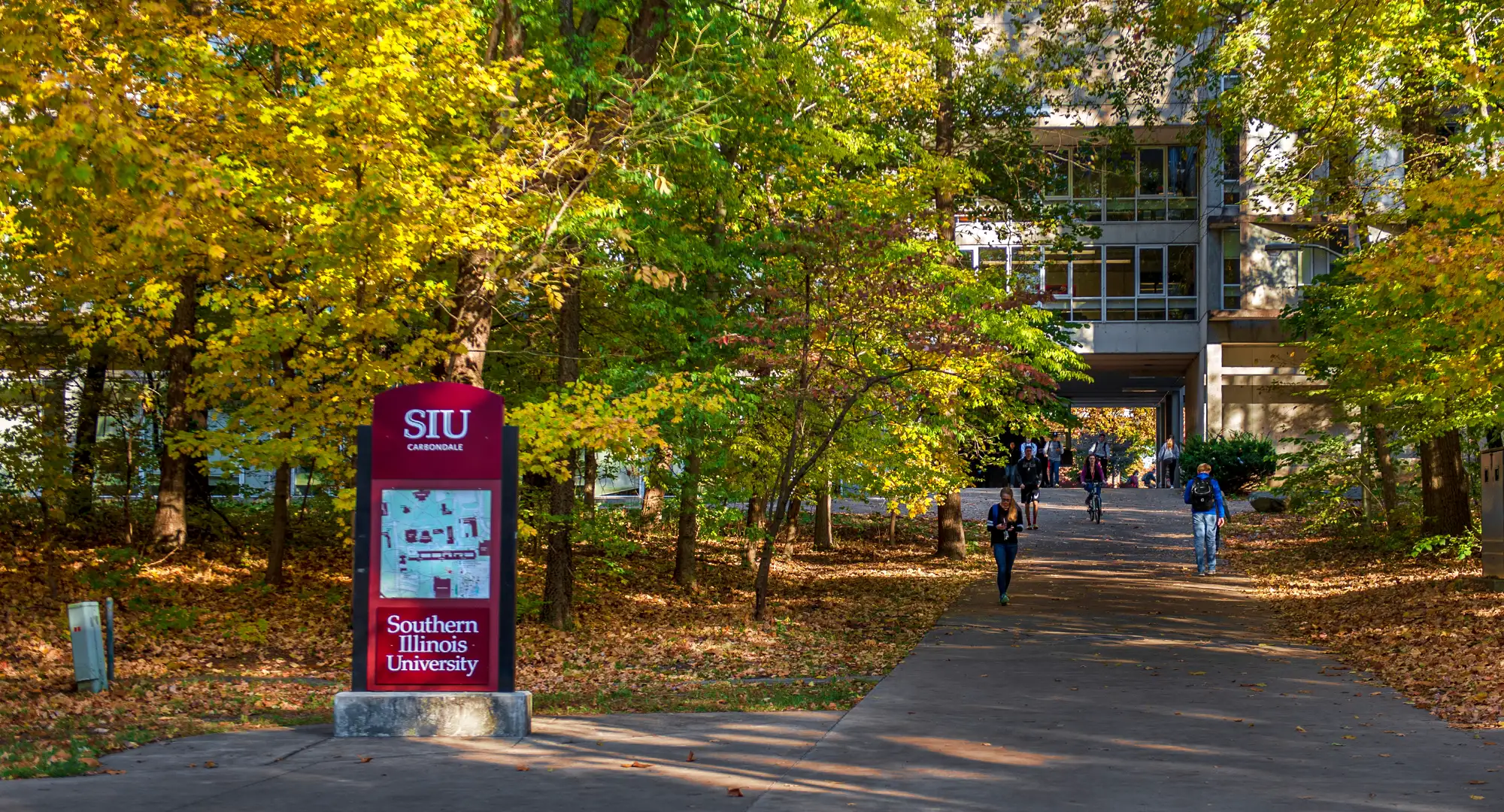 Southern Illinois University (SIU) campus scene featuring a directory sign with the SIU logo and 'Southern Illinois University' written on it, surrounded by trees with fall foliage. A pathway leads through the trees towards a building with students walking and biking along the path.