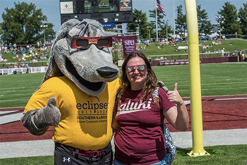 Grey Dawg and woman give the thumbs up while wearing eclipse glasses.