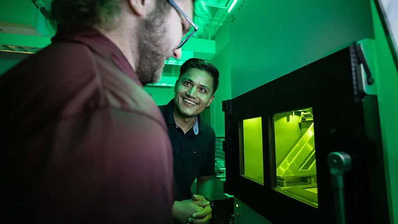 Bathed in a green light, two male graduate students chat while they stand next to a 3D printer.