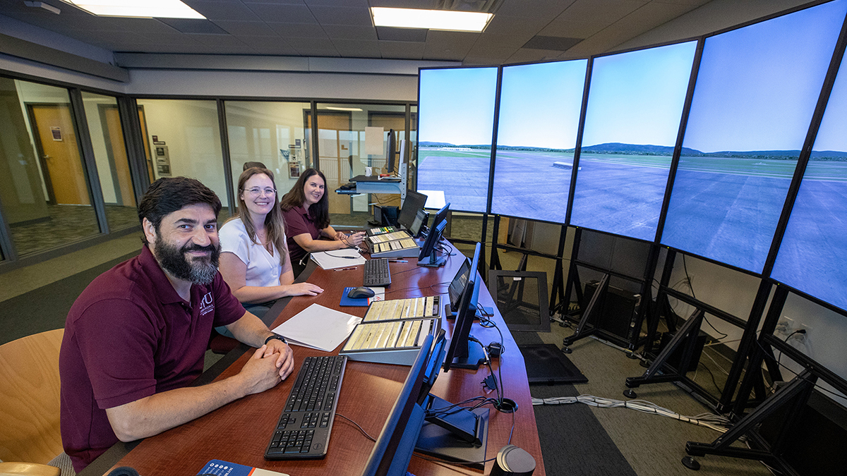 Three professors, one man and two women, sit a table in a simulated air traffic control tower, which includes large screens showing an outside view.