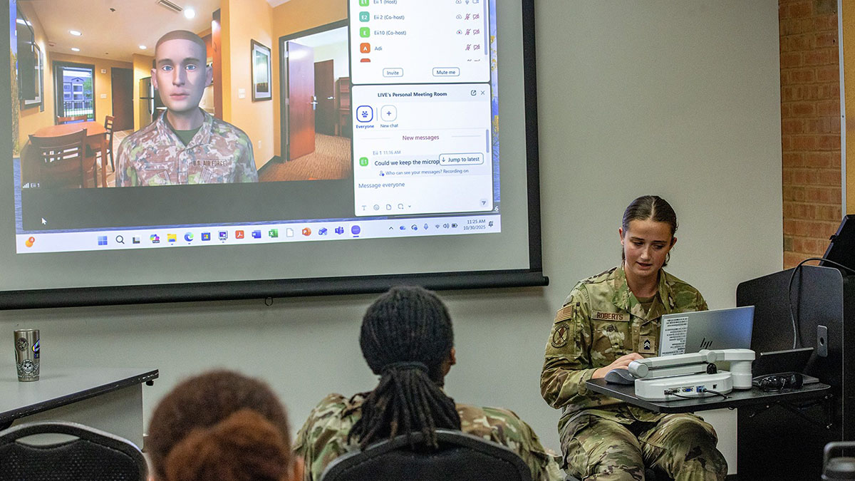 A young woman in camouflage interacts with a screen while a male avatar in camouflage gazes out at the class. 