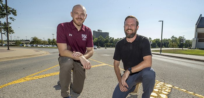 Brent Pease (left), associate professor of forestry and director of the newly established Roadside Ecology Center, and Ryan Campbell (right), director of the Center for Archaeological Investigations, kneel in a roadway near an entrance to the SIU campus on a bright, sunny day with a student housing dorm in the background.