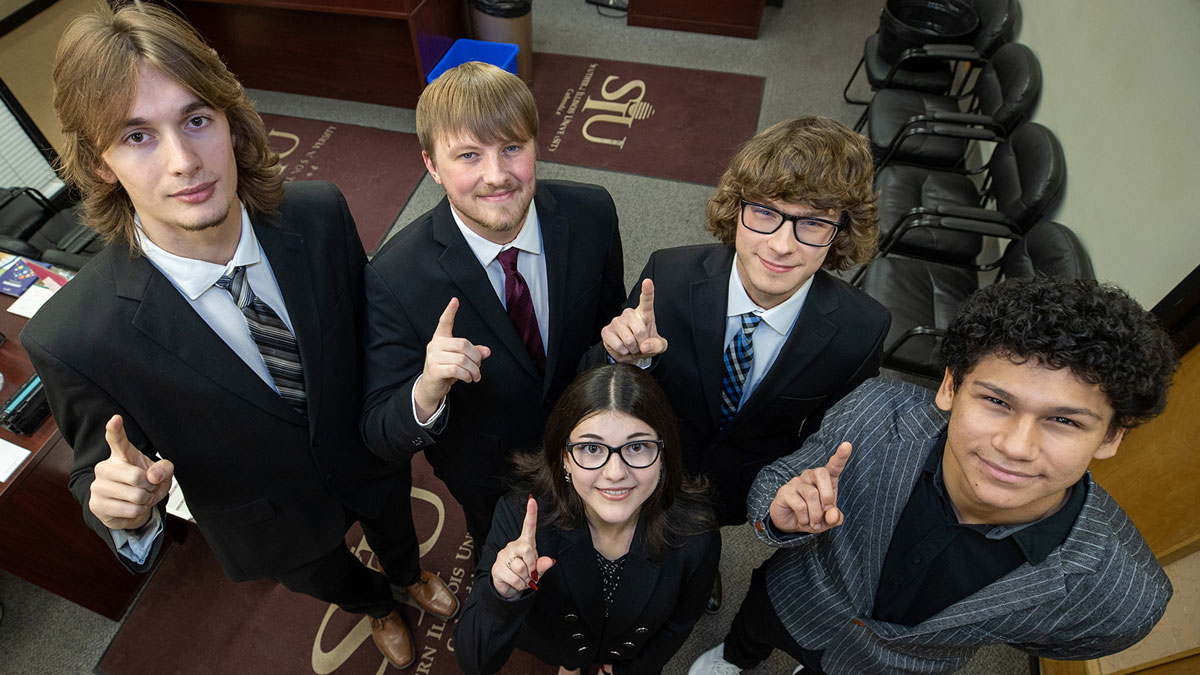 Professionally dressed students, four men and one woman, hold up their index fingers to indicate the team is number 1.
