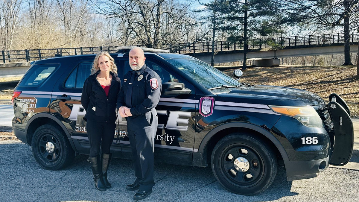 A counselor and a police officer stand next to a police car.