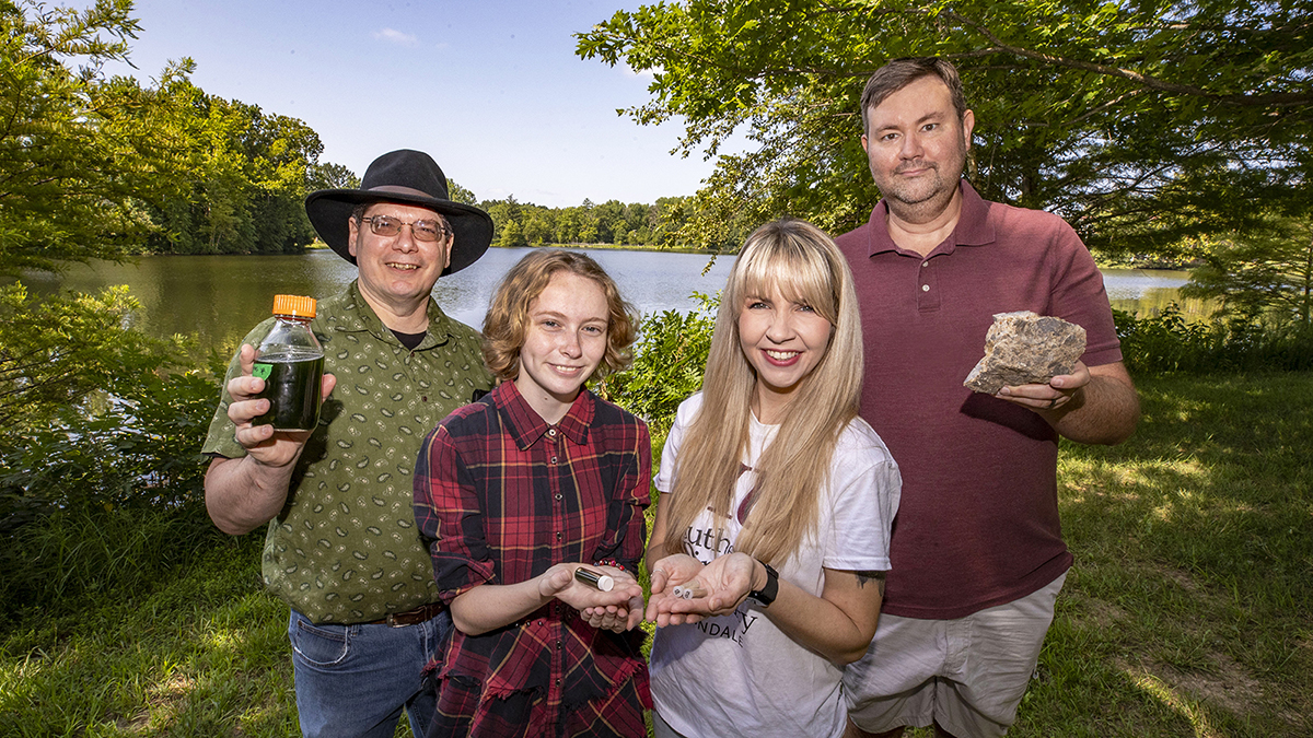 Four SIU researchers stand outside in front of Campus Lake, holding glass jars with algae and rock samples containing rare earth elements.