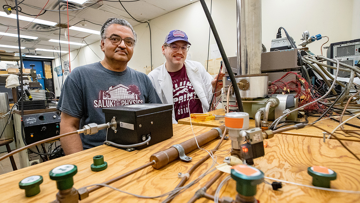 An older male professor and his younger male graduate student stand a table with lab equipment.