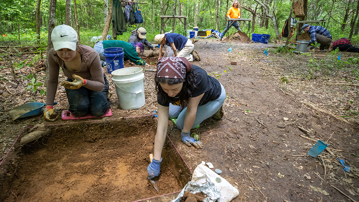 Kneeling archaeology students dig in a square plot while others work in the background.