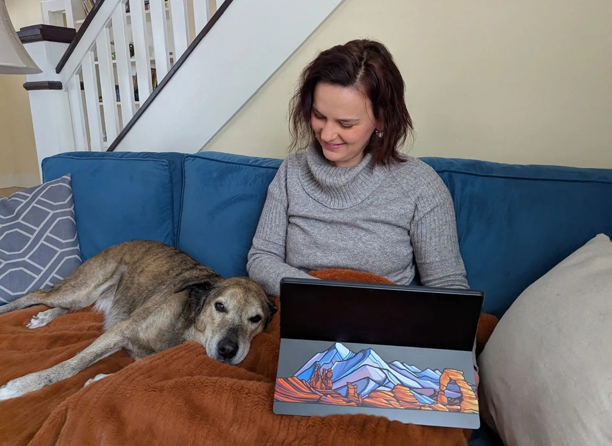Woman on couch working on laptop computer with dog laying on her lap.
