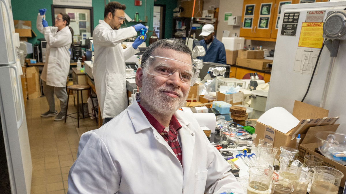 A professor works in a lab with students in the background.