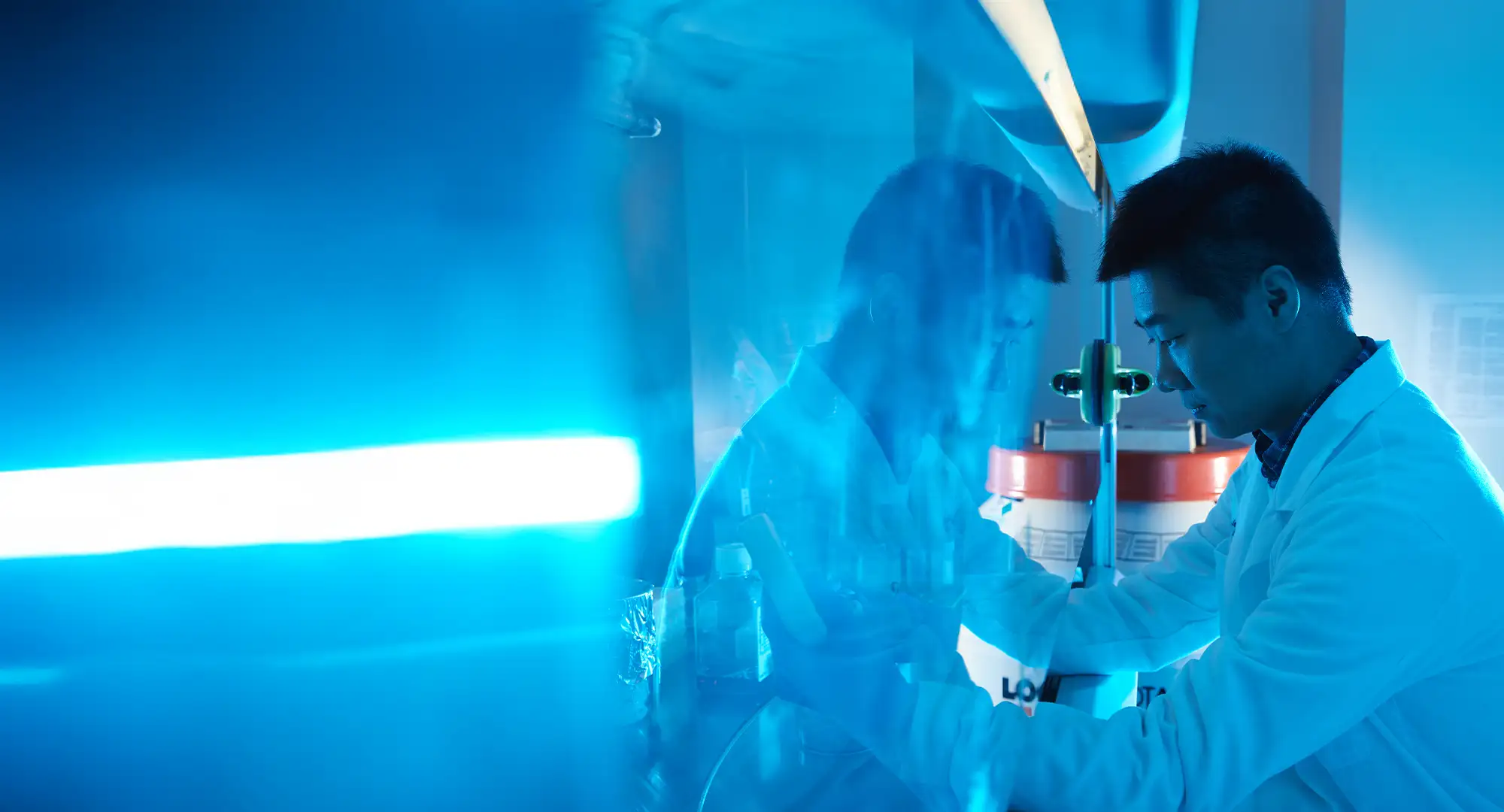 Xiangwei Fang in Nie's Research Lab, Med Microbiology. A scientist in a white lab coat working in a laboratory under blue lighting, with a reflection visible on a glass surface.