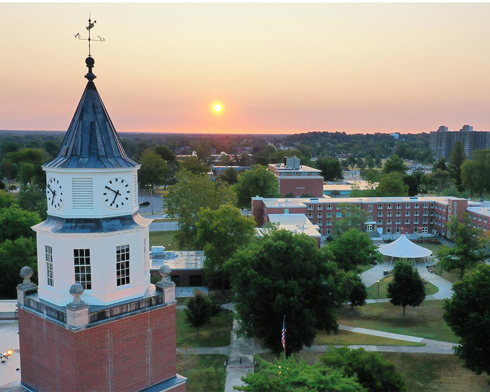 Pulliam clock tower on Southern Illinois University campus at sunset, with surrounding buildings, trees, and a pink-orange sky in the background.