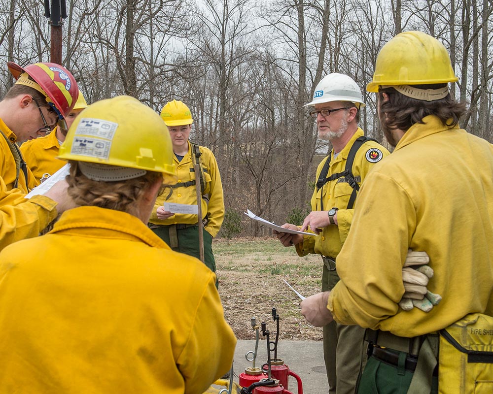 A group of individuals in yellow firefighter gear gathered outdoors, listening to a man holding a paper and wearing a white hard hat.