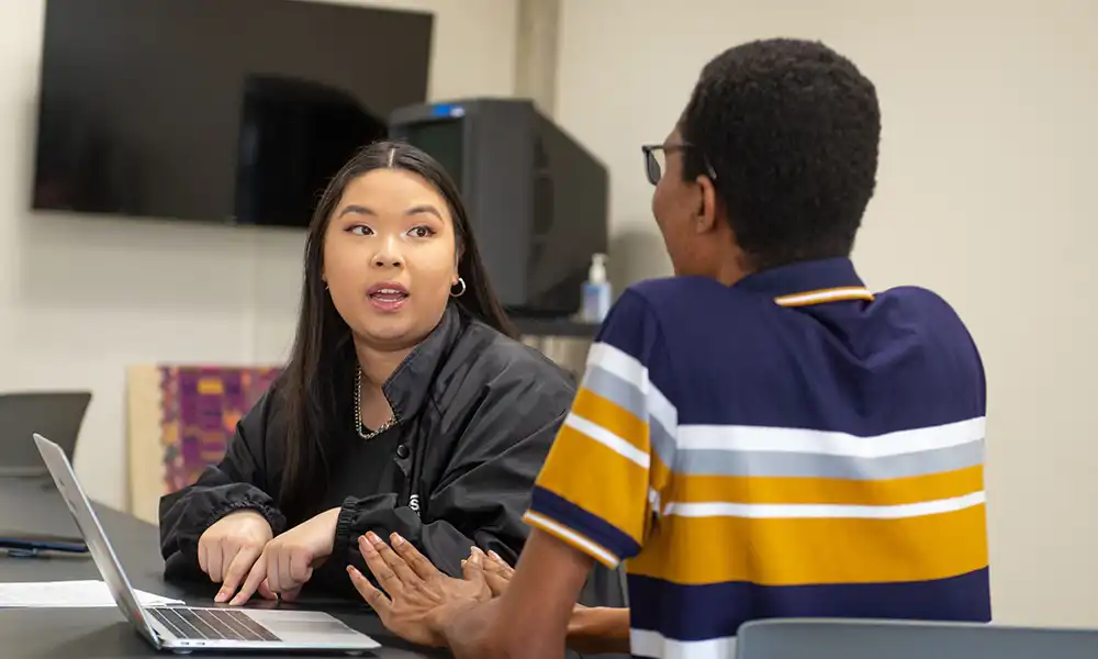 A student sits with an advisor at a table, talking while a laptop is open between them.