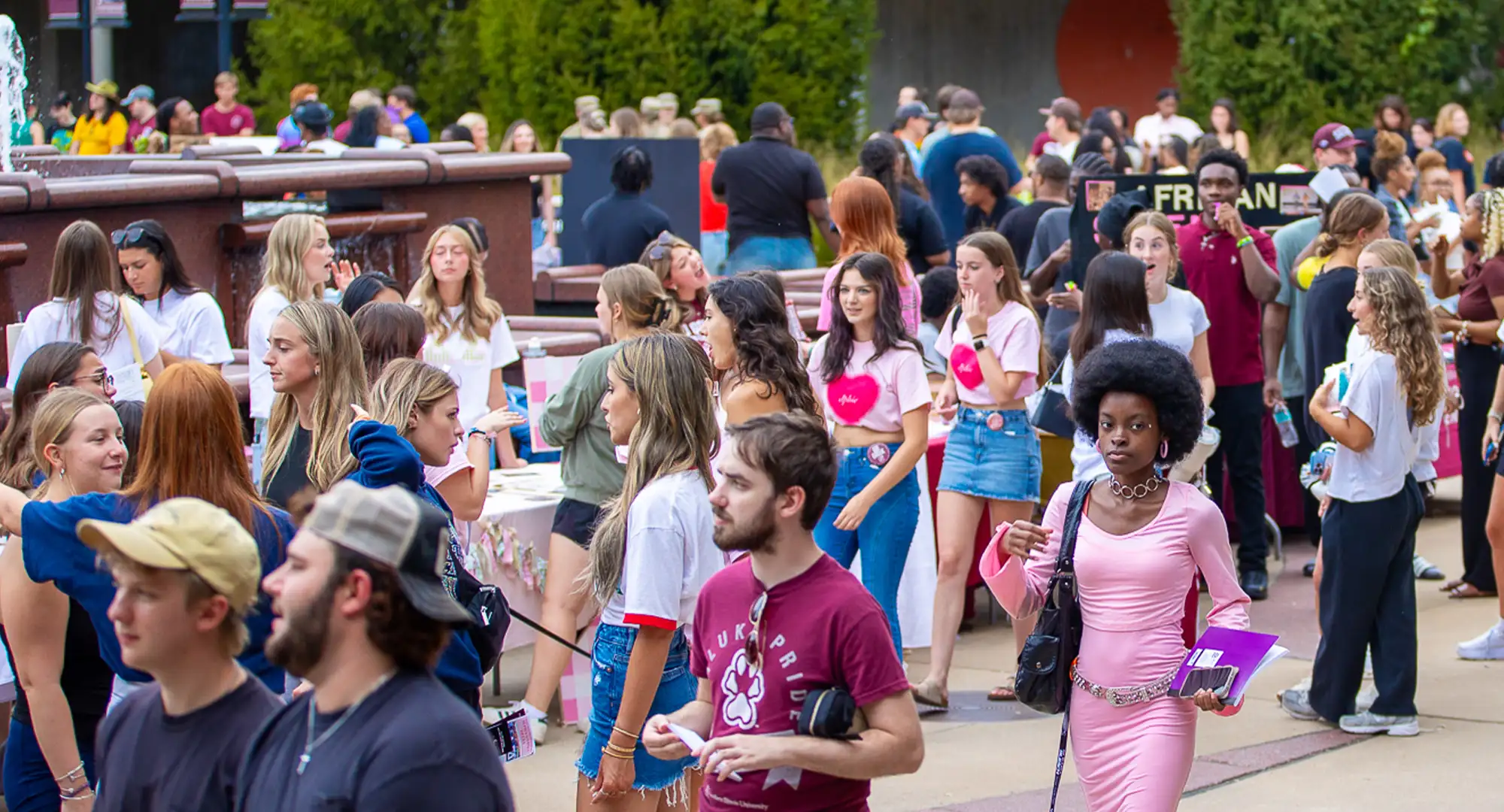 A large crowd of students walk and gather around tables at an outdoor campus event near a fountain.