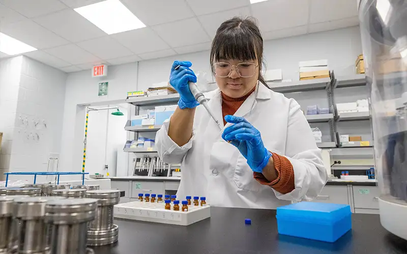 Student in a white lab coat and blue gloves uses a pipette to transfer liquid into vials during a laboratory experiment at Southern Illinois University.