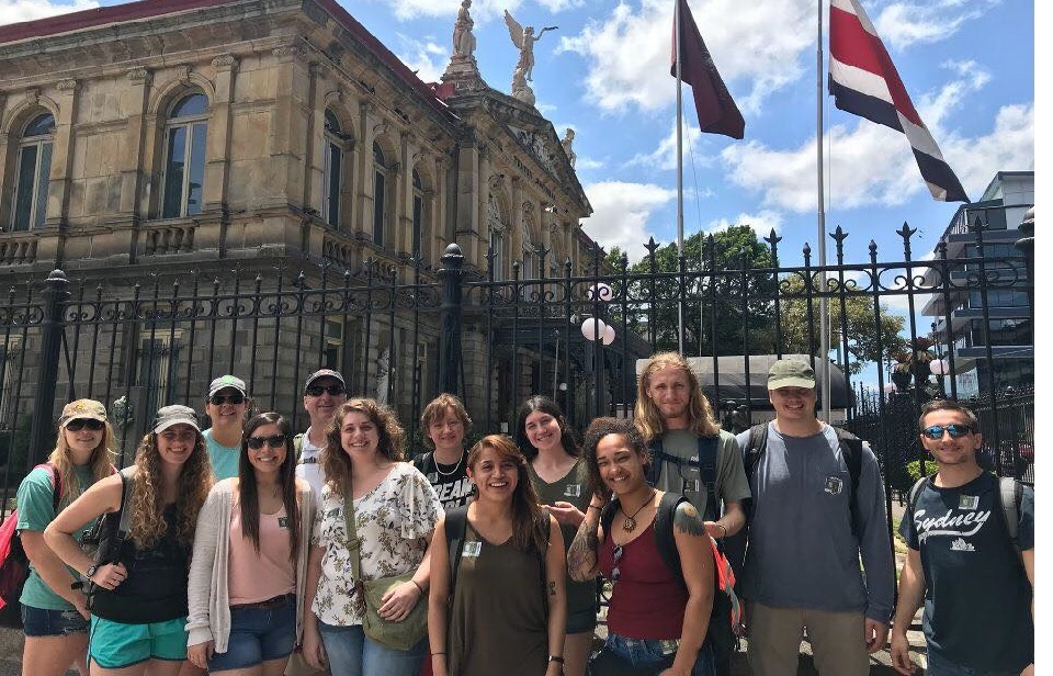 Group of students on a study abroad experience, smiling in front of a historic building in Costa Rica with flags waving above.
