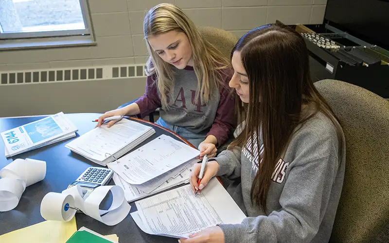 Two students review financial documents together at a desk, using calculators and tax forms during a budgeting or accounting exercise.