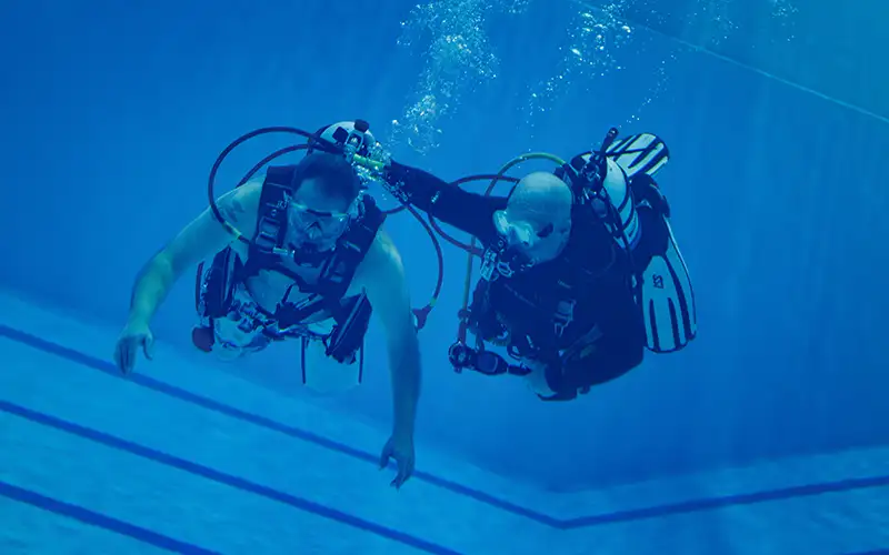 Instructor and student practice scuba diving techniques underwater in a swimming pool, both wearing full scuba gear.