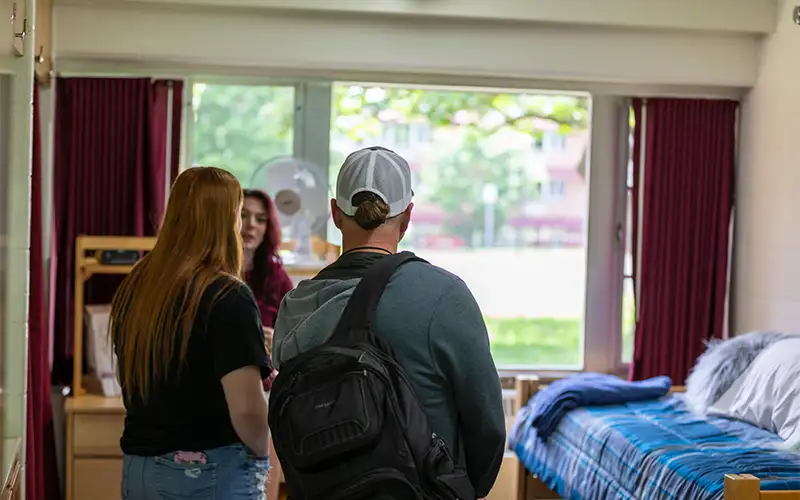 Students and a guide tour a campus residence hall room at Southern Illinois University, with a made bed, desk, and large window providing natural light.