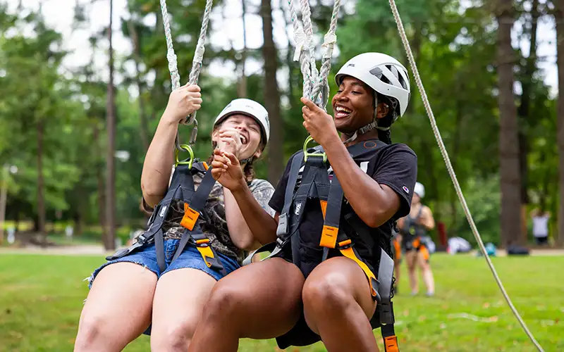 Two students wearing helmets and harnesses laugh together while suspended on a rope swing during an outdoor adventure activity.
